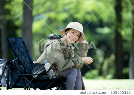 一位微笑的女子獨自一人在鬱鬱蔥蔥的森林中露營,她放鬆地坐在露營椅上,直視鏡頭。 一位微笑的女子獨自一人在鬱鬱蔥蔥的森林中露營,她放鬆地坐在露營椅上,直視鏡頭。 138955322