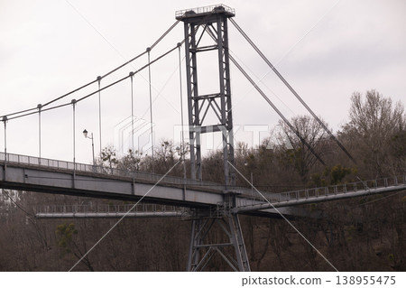 Large Gray Metal Suspension Bridge Structure Against Overcast Sky and Winter Trees Large Gray Metal Suspension Bridge Structure Against Overcast Sky and Winter Trees 138955475