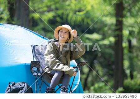 A smiling woman relaxes in a camping chair amidst the lush green forest, enjoying the scenery during her solo camping trip. A smiling woman relaxes in a camping chair amidst the lush green forest, enjoying the scenery during her solo camping trip. 138955490