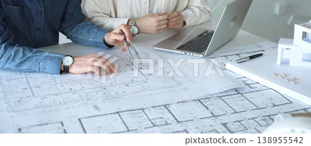 Architects engaging in a professional meeting, reviewing detailed blueprints on a table, utilizing a laptop, and collaborating on building design and construction planning 138955542