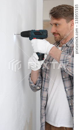 Middle aged male professional worker wearing grey checked shirt and white gloves, is drilling hole in a wall with cordless drill during home renovation work. Vertical portrait view Middle aged male professional worker wearing grey checked shirt and white gloves, is drilling hole in a wall with cordless drill during home renovation work. Vertical portrait view 138955585