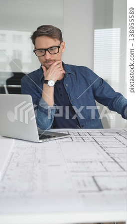 Male architect in glasses at a contemporary office working on a white architectural scale model of a modern house design, developing creative housing plans on a desk with blueprints and a laptop Male architect in glasses at a contemporary office working on a white architectural scale model of a modern house design, developing creative housing plans on a desk with blueprints and a laptop 138955589