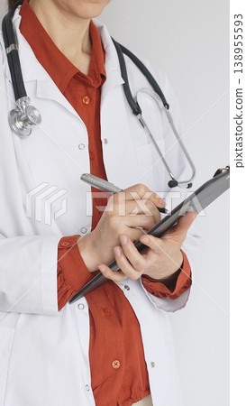 Close up of woman doctor wearing stethoscope and white coat over brick red blouse is standing and writing on a clipboard, representing medical expertise and patient care. Medicine concept Close up of woman doctor wearing stethoscope and white coat over brick red blouse is standing and writing on a clipboard, representing medical expertise and patient care. Medicine concept 138955593