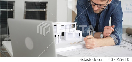 Male architect thinking while looking at a scale model of a modern house, developing architectural plans and blueprints for a new residential project in an office setting Male architect thinking while looking at a scale model of a modern house, developing architectural plans and blueprints for a new residential project in an office setting 138955618