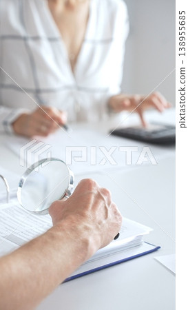 Close up of accountant hand using a magnifying glass and a calculators, to audit financial reports in brightly lit office. Audit and taxes concept 138955685
