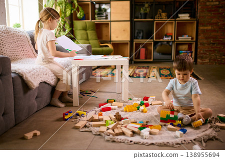 Little children playing separately at home with girl drawing and boy building blocks on floor. 138955694