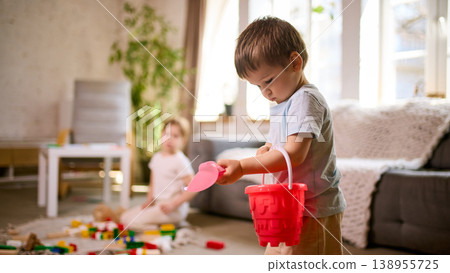 Little boy holding toy shovel and bucket while girl sits on floor playing with blocks. Little boy holding toy shovel and bucket while girl sits on floor playing with blocks. 138955725