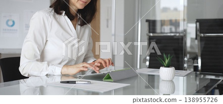 Businesswoman in white blouse and glasses sitting at a modern desk, working with a tablet in a contemporary office, focusing on digital tasks, using technology for efficient workflow Businesswoman in white blouse and glasses sitting at a modern desk, working with a tablet in a contemporary office, focusing on digital tasks, using technology for efficient workflow 138955726