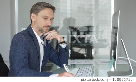 Businessman in a blue jacket thinking and brainstorming ideas while working on computer and taking notes in modern office. Business people concept Businessman in a blue jacket thinking and brainstorming ideas while working on computer and taking notes in modern office. Business people concept 138955733