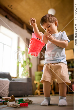 Little boy holding red bucket with toys standing in cozy home interior. Little boy holding red bucket with toys standing in cozy home interior. 138955734