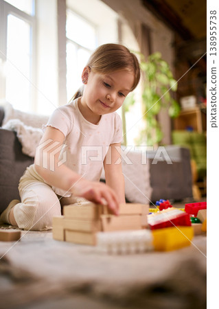 Little girl building wooden blocks structure while sitting on floor at home 138955738