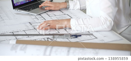 Architect's hands working on desk, comparing paper blueprints with digital floor plans on a laptop, using tools for precise measurements in architectural design Architect's hands working on desk, comparing paper blueprints with digital floor plans on a laptop, using tools for precise measurements in architectural design 138955859