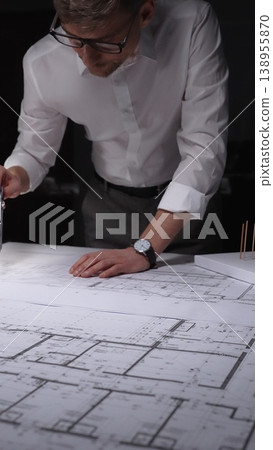 Architect working on blueprints using a compass, designing building plans in a studio at night. Concept of architecture and business in low key lighting Architect working on blueprints using a compass, designing building plans in a studio at night. Concept of architecture and business in low key lighting 138955870