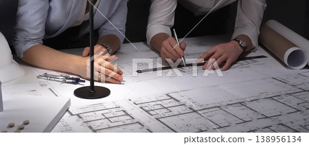 Architects collaborating on blueprints, diligently planning building design and construction project details late at night. Architects collaborating on blueprints, diligently planning building design and construction project details late at night. 138956134