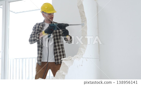 Male construction worker wearing beige checkered shirt, yellow hard hat and protective gloves, is demolishing white wall with rotary hammer drill, generating dust, medium long shot. Renovation Male construction worker wearing beige checkered shirt, yellow hard hat and protective gloves, is demolishing white wall with rotary hammer drill, generating dust, medium long shot. Renovation 138956141