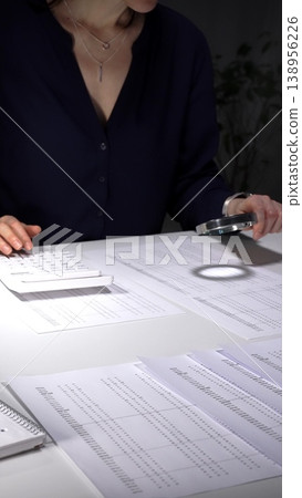 Person checking financial documents and numbers with a magnifying glass, analyzing business data for budgeting and taxes at night. Audit and taxes in business Person checking financial documents and numbers with a magnifying glass, analyzing business data for budgeting and taxes at night. Audit and taxes in business 138956226