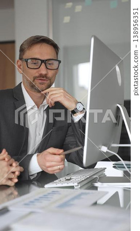 Businessman in a suit and glasses collaborating with a colleague during an office meeting, discussing business strategy while working at a modern computer desk, vertical view Businessman in a suit and glasses collaborating with a colleague during an office meeting, discussing business strategy while working at a modern computer desk, vertical view 138956351