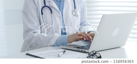 Female doctor documents patient information while working with digital devices at minimalist white workstation in medical office. Medicine and healthcare concept Female doctor documents patient information while working with digital devices at minimalist white workstation in medical office. Medicine and healthcare concept 138956363
