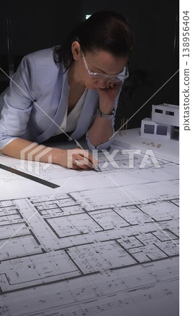 Female architect engrossed in work, reviewing blueprints and floor plans for a new construction project at night. Architecture and business concept in low key lighting Female architect engrossed in work, reviewing blueprints and floor plans for a new construction project at night. Architecture and business concept in low key lighting 138956404