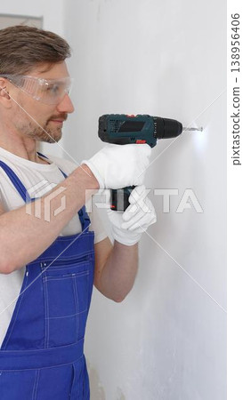Professional male construction worker in a blue jumpsuit, safety goggles, and work gloves is screwing a screw into the wall using a cordless screwdriver. Home renovation concept 138956406