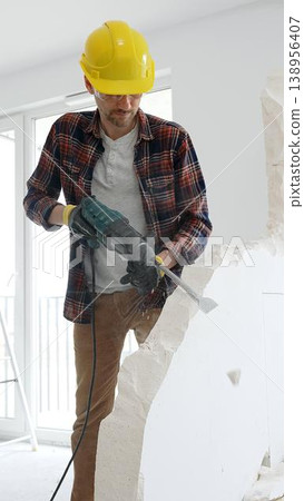 Man construction worker wearing red checkered shirt , yellow hard hat and protective gloves, is demolishing white wall with rotary hammer drill, generating dust, medium long shot. Renovation concept Man construction worker wearing red checkered shirt , yellow hard hat and protective gloves, is demolishing white wall with rotary hammer drill, generating dust, medium long shot. Renovation concept 138956407
