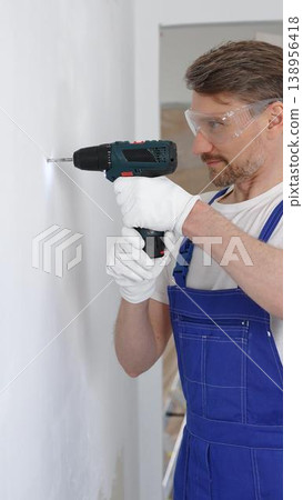 Professional male construction worker in a blue jumpsuit, safety goggles, and work gloves is screwing a screw into the wall using a cordless screwdriver, carrying out a careful home renovation 138956418
