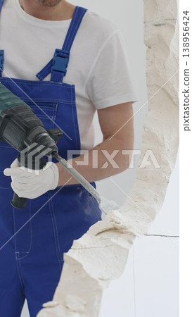 Unrecognizable male construction worker wearing blue overalls and protective white gloves, is demolishing white wall with rotary hammer drill, generating dust, close up horizontal view. Renovation Unrecognizable male construction worker wearing blue overalls and protective white gloves, is demolishing white wall with rotary hammer drill, generating dust, close up horizontal view. Renovation 138956424