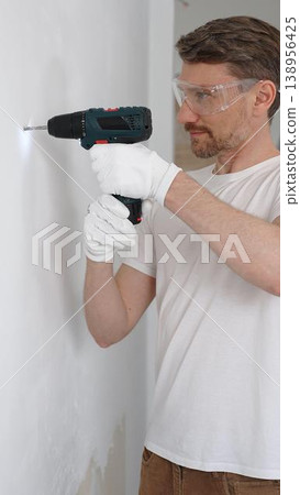 Middle aged male worker wearing white t-shirt and protective gloves, is drilling hole in a wall with cordless drill during home renovation work. Portrait view Middle aged male worker wearing white t-shirt and protective gloves, is drilling hole in a wall with cordless drill during home renovation work. Portrait view 138956425