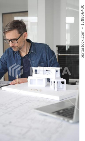 Male architect thinking intently while reviewing architectural blueprints and a 3d scale model of a contemporary house, brainstorming design ideas in a professional office environment 138956430