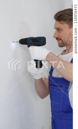 Man construction worker in a blue jumpsuit, safety goggles, and work gloves is screwing a screw into the wall using a cordless screwdriver, carrying out a careful home renovation. Vertical portrait 138956431