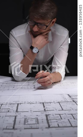 Architect analyzing blueprints late at night, a man wearing glasses thinking and concentrating on building plans. Concept of architecture and business in low key lighting Architect analyzing blueprints late at night, a man wearing glasses thinking and concentrating on building plans. Concept of architecture and business in low key lighting 138956485