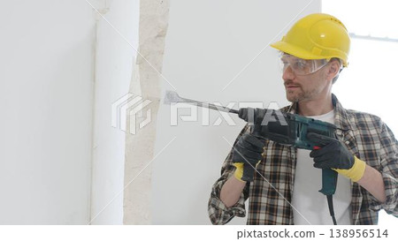 Portrait of a man construction worker wearing beige checkered shirt , yellow hard hat and protective gloves, is demolishing white wall with rotary hammer drill, generating dust. Renovation concept Portrait of a man construction worker wearing beige checkered shirt , yellow hard hat and protective gloves, is demolishing white wall with rotary hammer drill, generating dust. Renovation concept 138956514