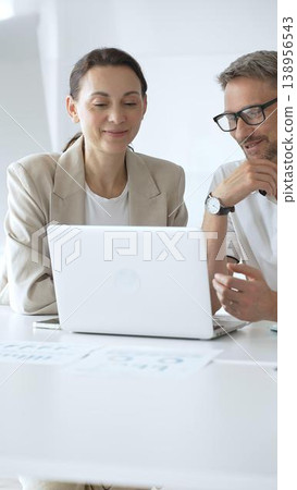 Businessman and adult businesswoman reviewing marketing analytics on laptops, working together strategically in contemporary workplace with minimalist interior design and collaborative atmosphere 138956543