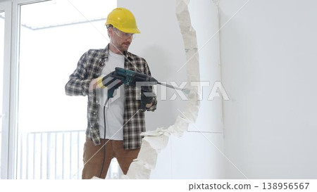 Male construction worker wearing beige checkered shirt, yellow hard hat and protective gloves, is demolishing white wall with rotary hammer drill, generating dust, medium long shot. Renovation project Male construction worker wearing beige checkered shirt, yellow hard hat and protective gloves, is demolishing white wall with rotary hammer drill, generating dust, medium long shot. Renovation project 138956567