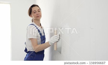 Woman construction worker wearing protective gloves and blue construction coveralls, spreading plaster smoothly across wall using professional drywall taping knife during renovation project. Portrait 138956687