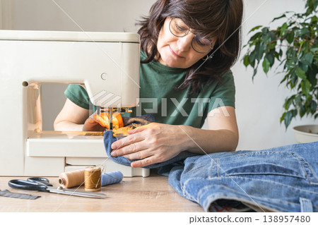 An adult woman repairs and alters denim jeans on a sewing machine at home, focused on an upcycling diy craft project. Reusing things. Environmental awareness. Eco-friendly lifestyle 138957480