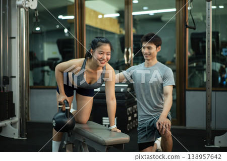 A woman is performing a kettlebell row on a bench while her trainer closely checks her form 138957642