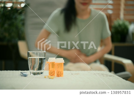 Pills and water on table while woman holds stomach in background Pills and water on table while woman holds stomach in background 138957831