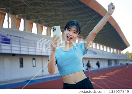 Asian woman celebrating fitness progress while checking smartphone on outdoor running tracking application 138957849