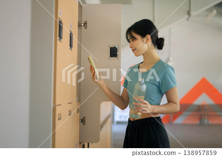Woman checking smartphone while holding water bottle at gym locker after workout, fitness routine, recovery moment and healthy lifestyle 138957859
