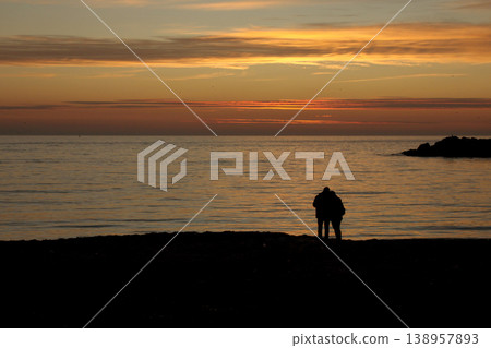 Silhouette of Couple on Beach at Sunset. A peaceful scene of a couple enjoying a golden sunset by the calm sea, captured in silhouette on a sandy shore. Silhouette of Couple on Beach at Sunset. A peaceful scene of a couple enjoying a golden sunset by the calm sea, captured in silhouette on a sandy shore. 138957893
