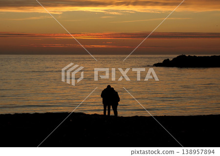 Silhouette of Couple on Beach at Sunset. A peaceful scene of a couple enjoying a golden sunset by the calm sea, captured in silhouette on a sandy shore. Silhouette of Couple on Beach at Sunset. A peaceful scene of a couple enjoying a golden sunset by the calm sea, captured in silhouette on a sandy shore. 138957894