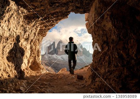 Male tourist walking in Grotta delle Tre Cime rocky tunnel framing sunset view of Tre Cime di Lavaredo mountain peak in Dolomites, Italy Male tourist walking in Grotta delle Tre Cime rocky tunnel framing sunset view of Tre Cime di Lavaredo mountain peak in Dolomites, Italy 138958089
