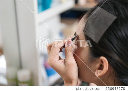 Beautiful Thai woman applying makeup with eye liner, eyebrow pencil in front of mirror. Lifestyle concept for cosmetics and self care 138958278