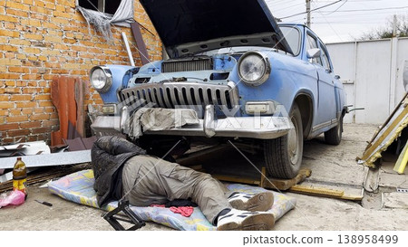 Male repairman lies under his vintage car at courtyard of house. Auto mechanic repairing his blue retro vehicle outdoor. Concept of automobile maintenance. Slow motion 138958499