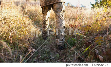 Low view to male feet of ukrainian soldiers going along trail at countryside. Legs of young army man walking through dry grass at sunny day. War in Ukraine. Slow motion Low view to male feet of ukrainian soldiers going along trail at countryside. Legs of young army man walking through dry grass at sunny day. War in Ukraine. Slow motion 138958523