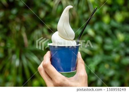 A soft-serve ice cream cone in a hand against a lush green natural background. A soft-serve ice cream cone in a hand against a lush green natural background. 138959249