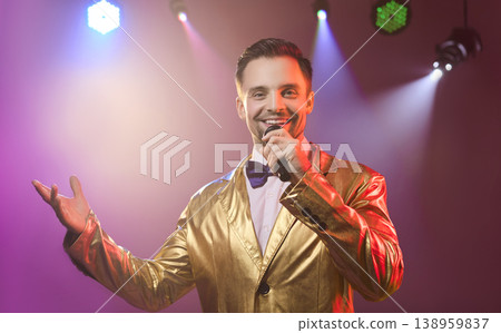 Smiling showman in gold suit singing with microphone under disco lights on stage Smiling showman in gold suit singing with microphone under disco lights on stage 138959837