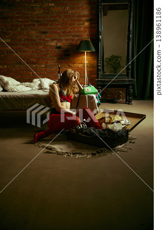 Woman sitting by open suitcase with phone in hand in moody loft bedroom interior. 138961186
