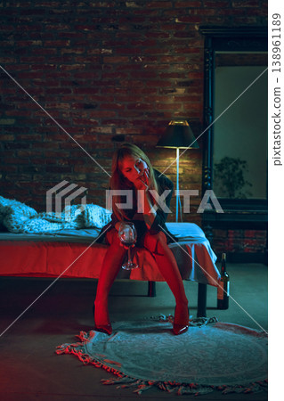 Young woman sitting on bed with glass of wine in dramatic red blue lighting moody interior. Young woman sitting on bed with glass of wine in dramatic red blue lighting moody interior. 138961189